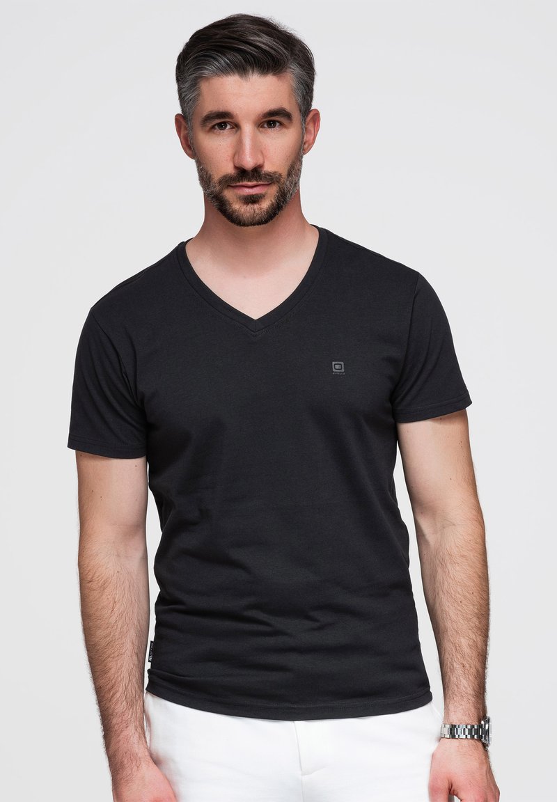 Homme aux cheveux courts et foncés et à la barbe, debout face à l'avant, portant un t-shirt noir à col en V et un pantalon blanc sur un fond uni.