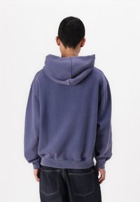 Man i lila huvtröja och mörkblå jeans vänd bort mot en enkel vit bakgrund.