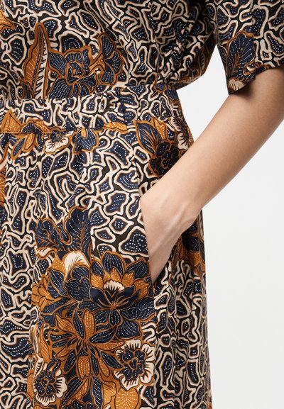 Χέρι που ξεκουράζεται στην τσέπη φορέματος με λουλουδάτο σχέδιο σε μαύρα, καφέ και κρεμ χρώματα με περίπλοκα σχέδια φύλλων και λουλουδιών.