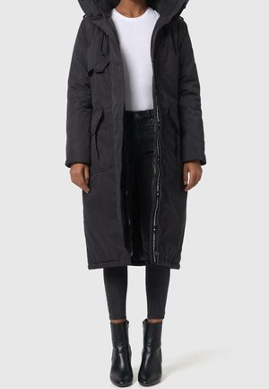 Person iført en lang sort hættetrøje vinterfrakke over en hvid skjorte, sort stramme jeans og sorte ankelstøvler, stående foran en ensfarvet baggrund.