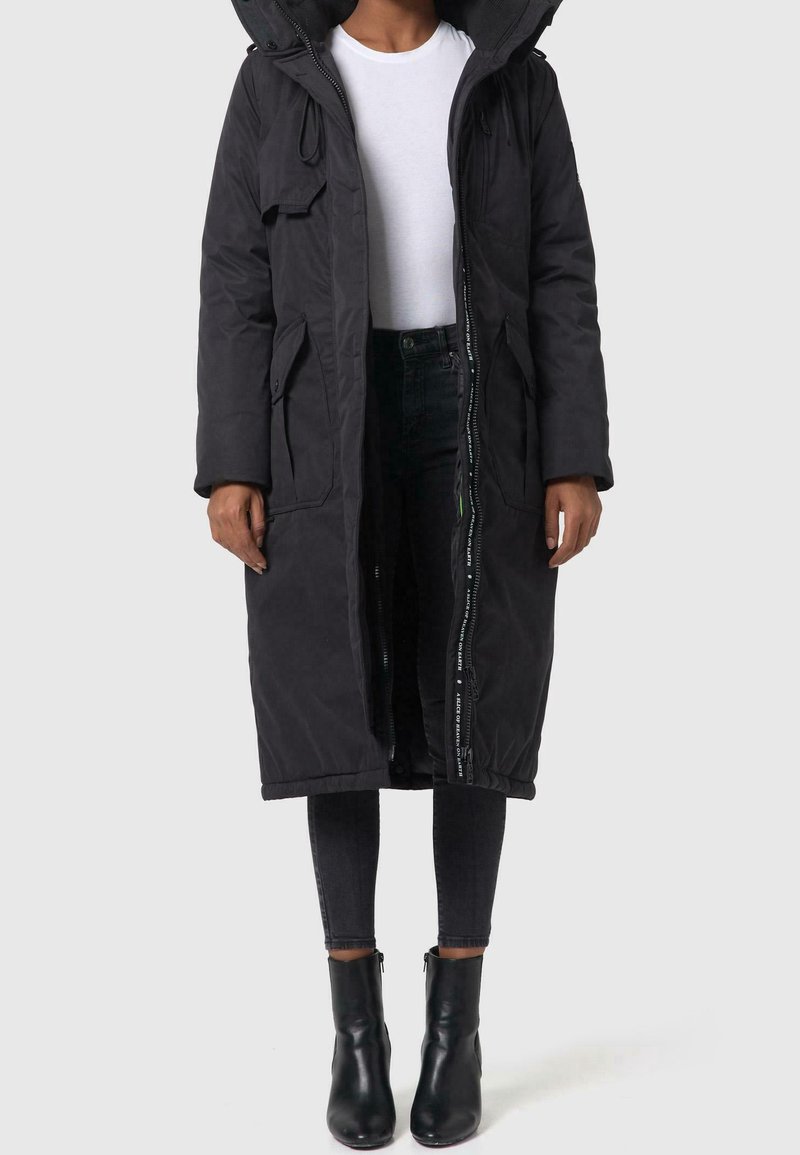 Personne portant un long manteau d'hiver noir à capuche par-dessus une chemise blanche, un jean skinny noir et des bottines noires, debout sur un fond uni.