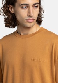 Jonge man met gemiddeld lang golvend haar, gekleed in een eenvoudige bruine T-shirt met een klein geborduurd logo op de borst, die opzij kijkt.