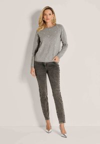 Frau in einem hellgrauen Pullover mit kleinen Verzierungen, dunklen grauen Slim-Fit-Jeans und silbernen, spitzen Absätzen, vor einem einfarbigen Hintergrund stehend.