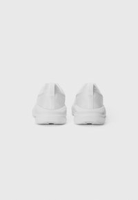 Une paire de chaussures de sport blanches vues de l'arrière, avec un tissu texturé et des semelles épaisses, sur un fond blanc uni.