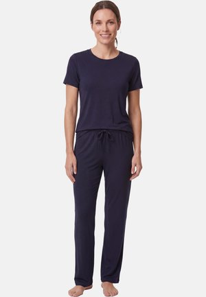 Vrouw staat blootsvoets, draagt een effen donkerblauw T-shirt met korte mouwen en bijpassende broek met koordje, glimlacht met haar naar achteren gebonden.
