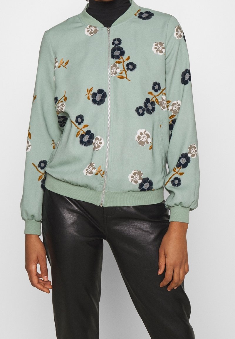 Mintgrüner Bomberjacke mit floralen Motiven in Weiß, Marineblau und Braun. Hat einen Reißverschluss vorne, gerippte Manschetten und Saum sowie eine glatte Textur.