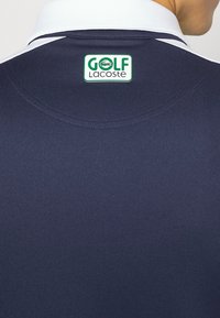 Marinblå golfpolo med vit krage, med en grön och vit logotyp på baksidan som står "GOLF Lacoste." Slät textur.