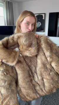 Jeune femme aux cheveux blonds tenant un grand manteau en fourrure marron à l'intérieur, debout dans une pièce avec des œuvres d'art encadrées et des rideaux en arrière-plan.