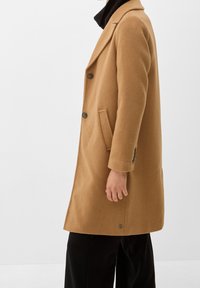 Persona che indossa un cappotto di lana color cammello con bottoni e tasche laterali, abbinato a pantaloni neri e un dolcevita nero.