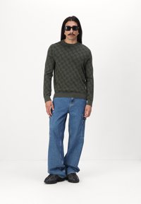 Grüner Strickpullover mit einem strukturierten Muster, kombiniert mit hellblauen weit geschnittenen Jeans und schwarzen Sneakers. Sonnenbrillen runden den Look ab.