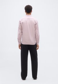 Άνδρας όρθιος με την πλάτη προς το μέρος του θεατή, φοράει ανοιχτό ροζ πουκάμισο μακρυμάνικο και μαύρο παντελόνι, μπροστά σε απλό λευκό φόντο.