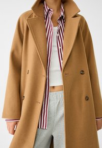 Manteau en laine marron à double boutonnage, grands revers et deux boutons. Porte par-dessus une chemise rayée rouge et blanche et un pantalon de survêtement gris.