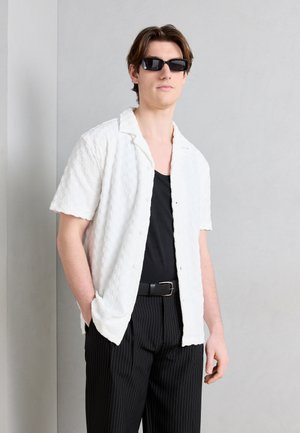 Junger Mann mit schwarzen Sonnenbrillen, weißem, strukturiertem Kurzarm-Hemd mit Knöpfen, schwarzem Tanktop und schwarzen Nadelstreifenhosen, die Hände in den Taschen.