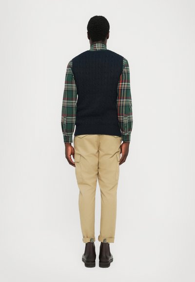 Hombre de pie de espaldas, vistiendo un chaleco de punto oscuro, camisa a cuadros verde y roja, pantalones cargo beige y botas marrón oscuro.