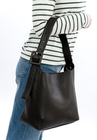Persoon die een blauwe jeans en een gestreept shirt draagt, draagt een grote zwarte leren schoudertas met een verstelbare gespband.