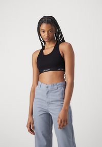 Black crop top with a wide round neckline, featuring a branded elastic band. Pair of light blue high-waisted pants with a smooth texture.