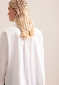 Biała bluzka z plisą z tyłu, długimi rękawami i miękką teksturą. Czyste linie i nieco luźniejszy krój. Minimalistyczny design.