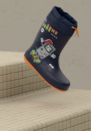 Bota de lluvia infantil azul marino con suela naranja, parte superior con cordón y gráfico de una consola de videojuegos montando en patineta.
