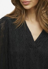 Blusa nera in tessuto testurizzato con scollatura a V e intricate fantasie a onda, con accenti trasparenti sulle maniche. Dettaglio in tessuto morbido visibile.