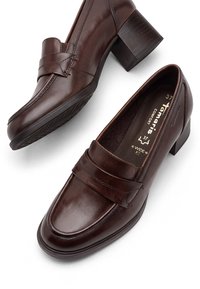 Paire de mocassins en cuir marron foncé à talon moyen, largeur large, bout carré et bride décorative sur le dessus, nom de marque visible à l'intérieur d'une chaussure.
