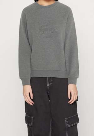 Personne portant un sweat Lacoste gris avec logo embossé et un pantalon cargo noir avec des coutures blanches, debout les mains détendues.