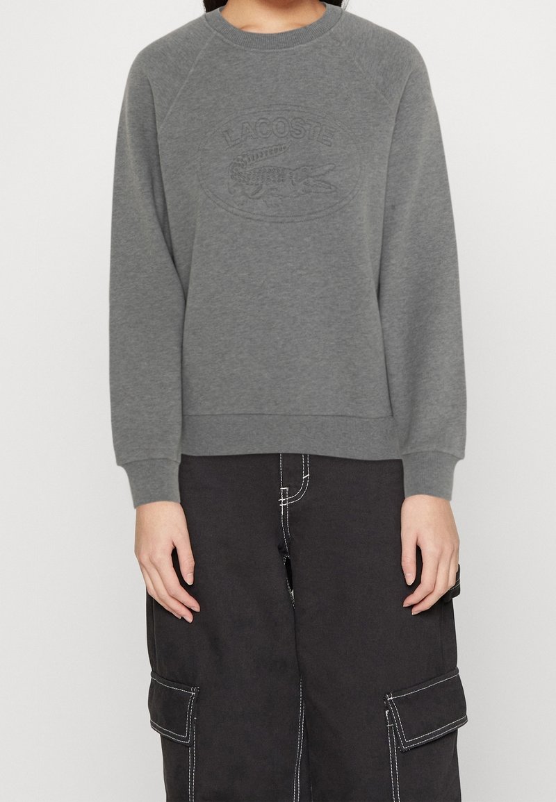 Personne portant un sweat Lacoste gris avec logo embossé et un pantalon cargo noir avec des coutures blanches, debout les mains détendues.