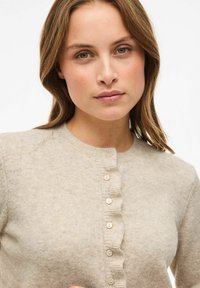 Bež pletena jakna iz lahkega materiala z okroglo vratno odprtino, rebrastim gumbom in petimi kremnimi gumbi. Mehak otip z diskretnimi madeži.