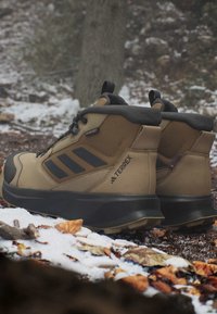 Paar braune und schwarze Adidas Terrex Wanderschuhe, die auf verschneitem Waldboden mit gefallenem Laub und Baumstamm im Hintergrund ruhen.