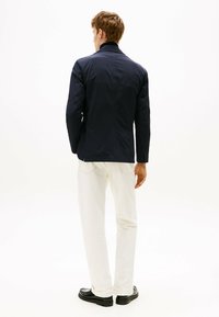 Giacca blu navy con colletto, dal design strutturato e texture liscia, abbinata a pantaloni bianchi e scarpe nere.