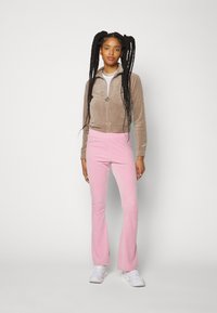 Burgunderfarbene Samt-Zip-Jacke mit hohem Kragen, kombiniert mit rosa ausgestellten Hosen. Das Model trägt weiße Sneakers; der Hintergrund ist reinweiß.