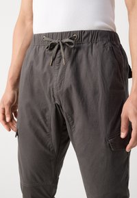 Pantalon cargo gris avec une taille élastique et un cordon de serrage. Ils comportent des poches latérales et une texture de tissu lisse et léger.