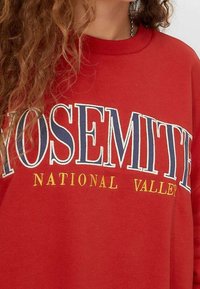 Rode sweatshirt met "YOSEMITE" in grote, witte en blauwe letters, met "NATIONAL" in oranje en "VALLEY" eronder in een kleiner lettertype.