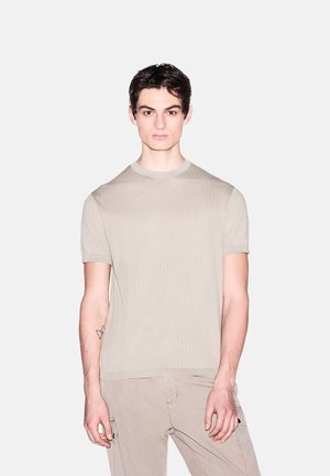 Jonge man met donker haar, gekleed in een beige gebreid shirt met korte mouwen en lichtbeige cargobroek, tegen een effen grijze achtergrond.