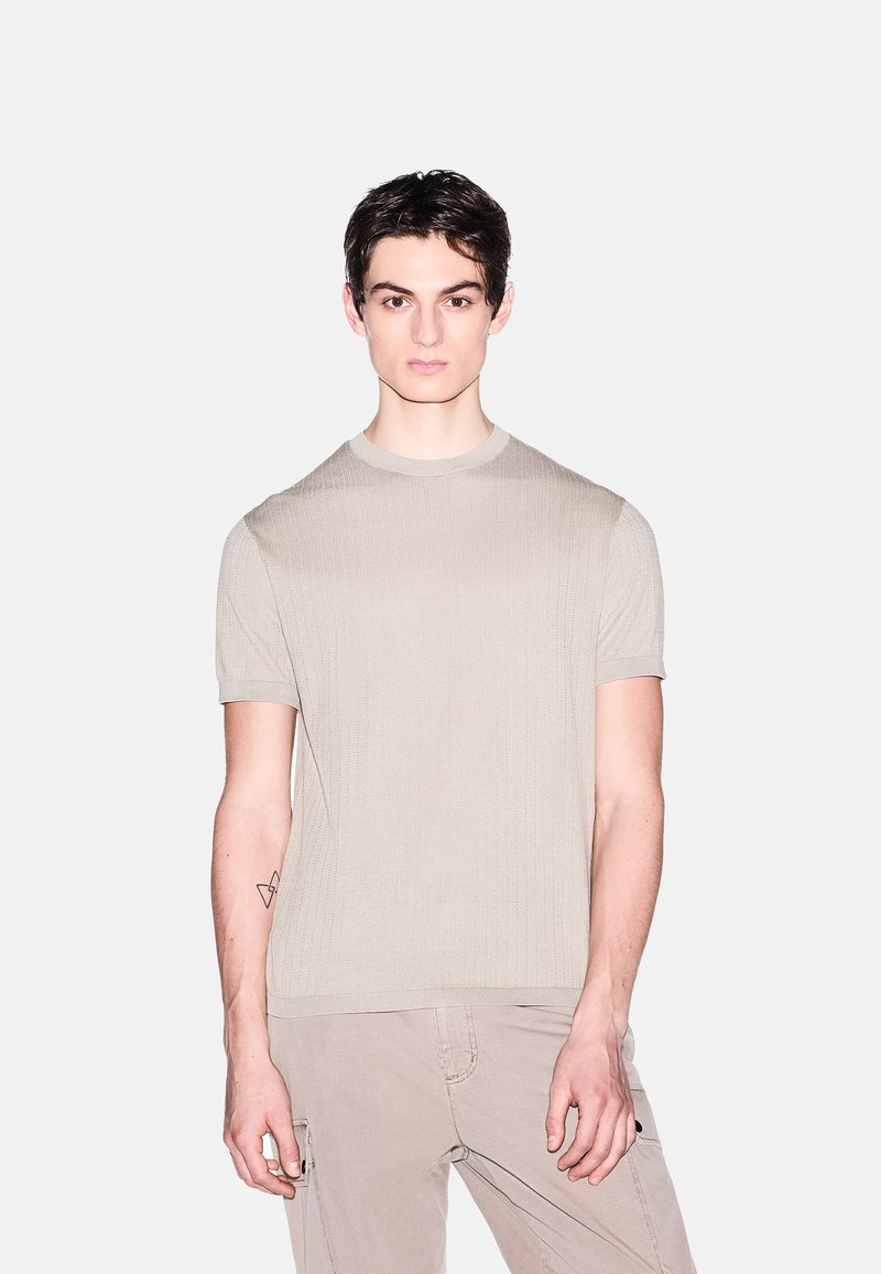 Jeune homme aux cheveux foncés portant une chemise en tricot beige à manches courtes et un pantalon cargo beige clair sur un fond gris uni.