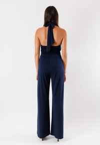 In tuta blu navy con corpino arricciato e collo all'americana. Pantaloni a gamba ampia e dettaglio con nodo al collo, realizzato in tessuto morbido e fluido.