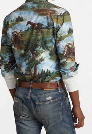 Koszula z długim rękawem z motywami wiejskiej jazdy konnej, drzewami i napisem, noszona z brązowym paskiem i postarzałymi niebieskimi dżinsami.