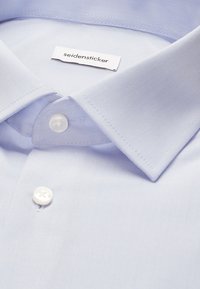 Col et boutons d'une chemise bleu clair avec une étiquette blanche visible indiquant le nom de la marque seidensticker.