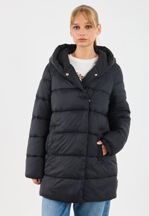 Jeune femme portant une veste rembourrée noire à capuche avec les mains dans les poches, un t-shirt graphique blanc et un jean bleu, se tenant contre un fond blanc.