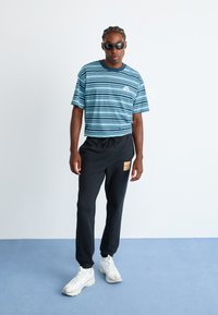 Mann mit geflochtenem Haar, der schwarze Sonnenbrillen, ein blau gestreiftes Nike-T-Shirt, schwarze Columbia-Sweatpants und weiße Sneakers trägt, steht auf einem blauen Boden.