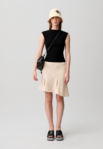 Giovane donna in piedi con top nero senza maniche, gonna beige, borsa nera a tracolla, cappello bianco a secchiello e sandali neri con plateau su sfondo chiaro.
