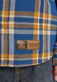 Modrá a žltá károvaná košeľa s koženou nášivkou, ktorá hovorí "DC Quality Goods Since 1994," nosená cez tmavomodré denimové džínsy.