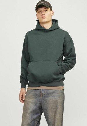 Felpa con cappuccio di colore verde scuro, in materiale cotone, dotata di una tasca frontale e polsini a coste, abbinata a jeans sbiaditi e a vestibilità morbida.