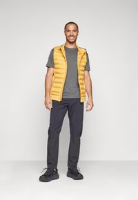 Chaleco amarillo acolchado sobre una camiseta gris, combinado con pantalones oscuros y zapatos negros. Textura suave y diseño ajustado con cierre de cremallera.