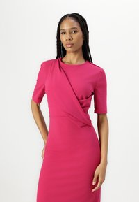 Robe ajustée à manches courtes en tissu rose vif avec un design torsadé à la taille et un col rond. Texture lisse, longueur au genou.