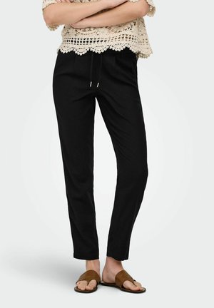 Nő, fekete, megkötős, rövidített nadrágban, bézs színű csipkefelsőben és barna, nyitott orrú szandálban, karját összefonva áll.