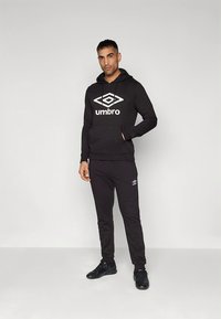 Svart hettegenser og joggebukser i mykt stoff, med en stor hvit Umbro-logo foran og subtil merkevare på buksene.