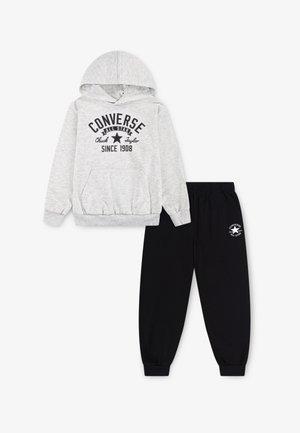 Sudadera gris con capucha, bolsillo frontal y estampado negro; pantalones de chándal negros con cintura elástica y logo blanco en el muslo izquierdo.