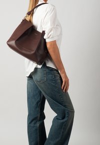 Bolso de hombro de cuero marrón con un diseño estructurado y relajado, detalles en metal dorado y una textura suave y lisa. Cuelga a la altura de la cadera.