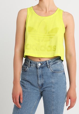 Henkilö, jolla on keltainen Adidasin lyhyt toppi ja korkeavyötäröiset siniset farkut yksiväristä taustaa vasten.