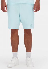 Pantalones cortos de color azul claro, hechos de una tela suave, con un corte relajado y un logotipo bordado en la parte inferior izquierda. Combinados con zapatos blancos.